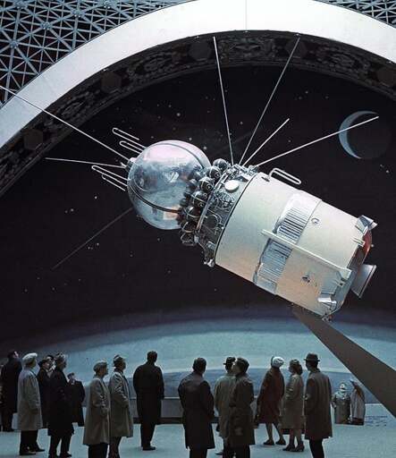 A group of people visiting an exhibit for a Vostok model, hanging overhead, in a wide exhibition room. 