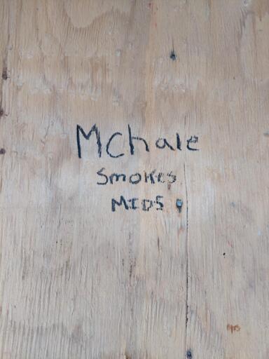 an unfinished plywood wall with "Mchale smokes mids" written in black marker with each word written on an individual line