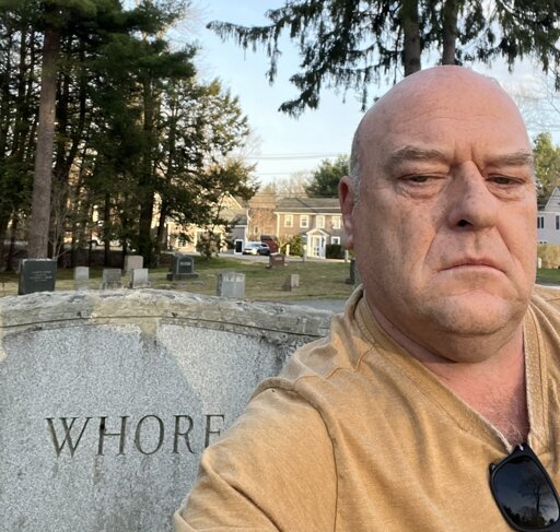 Dean Norris with his big bald head in front of a gravestone that reads "Whore". He has a very serious expression on his face and is wearing a yellow shirt.